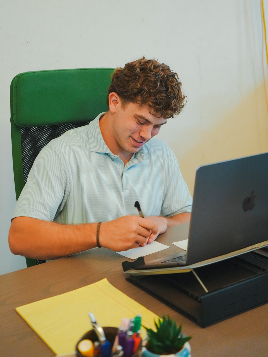 Marketing manager sitting at his desk writing on a note card with his laptop and monitor in front of him.