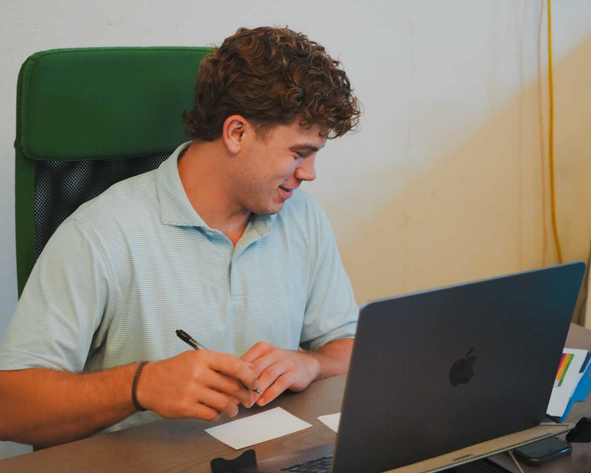 Marketing manager sitting at his desk writing on a note card with his laptop and monitor in front of him.